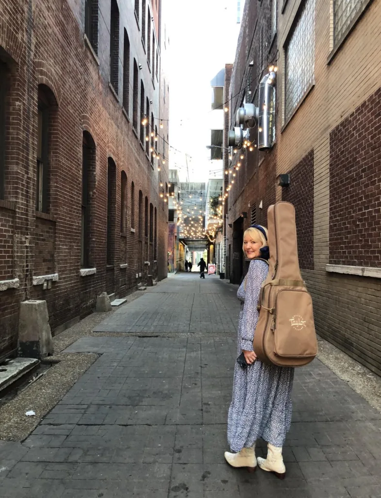 Stefani Morton standing in an alley in Nashville, TN. She has her guitar on her back and is looking over her shoulder.