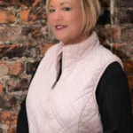 Stefani wearing a white vest over a black sweater standing in front of a brick wall.